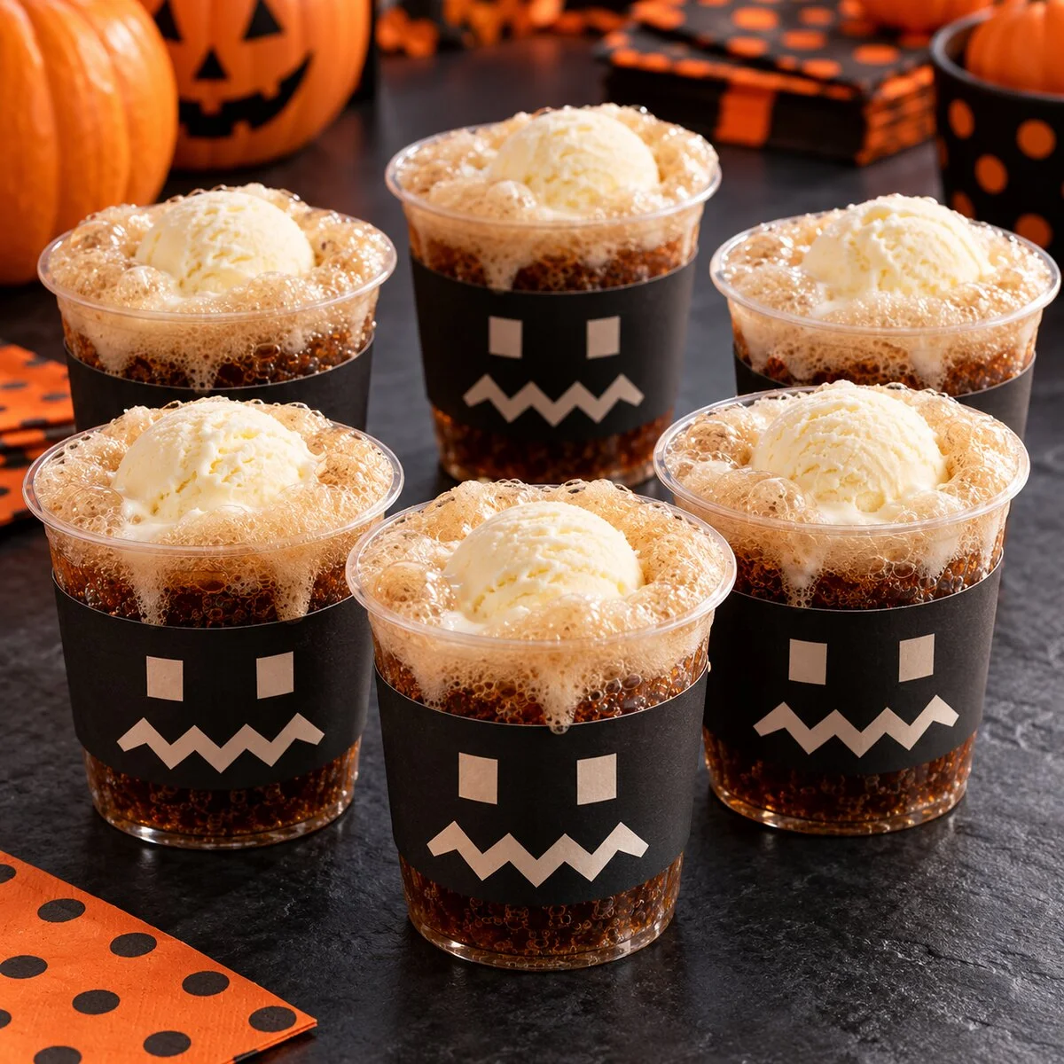 Six foaming root beer floats with vanilla ice cream in clear cups wrapped with black paper monster faces